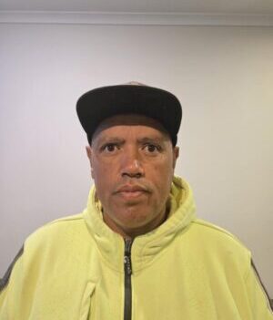Man wearing a yellow hoodie and black cap, facing the camera in a plain room.