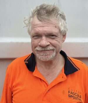 Portrait of an older man with gray hair and a goatee, wearing a bright orange work shirt, standing against a gray metal wall.