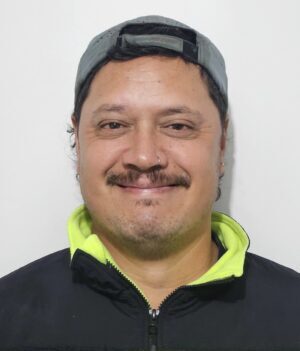 Smiling man wearing a gray cap and a neon yellow–black high-visibility jacket, looking at the camera.
