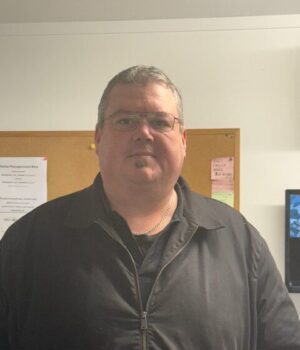 Front view of a middle-aged man with glasses and a black zip-up jacket, standing in an office with a bulletin board behind him.