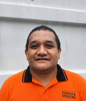 Portrait of a middle-aged man with short dark hair, wearing an orange polo shirt with a black collar, standing in front of a light gray wall.
