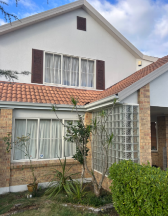 Two-story house with white walls, brown shutters, and a red-tiled roof; front garden with tall plants and a glass-block window feature.