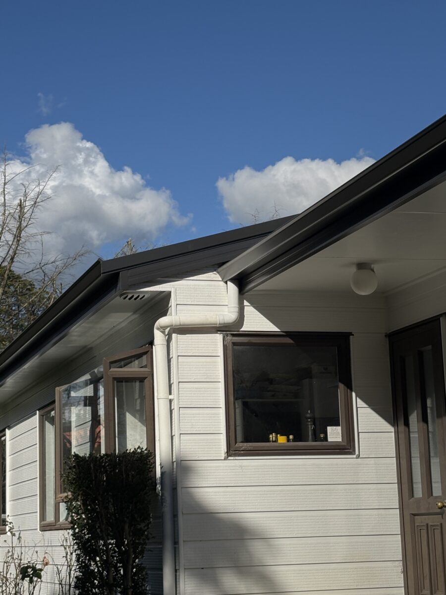 White house exterior with a slanted roof, white siding, a white downspout, and a large brown-framed window against a blue sky.