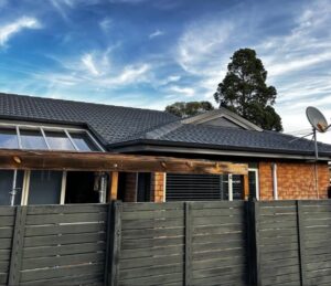 Backyard view of a brick house with a dark tiled roof, a wooden pergola, a tall privacy fence, and a satellite dish against a blue sky.