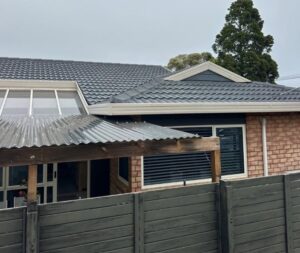 Exterior view of a single-story brick house with a metal tile roof, a corrugated carport, and a wooden panel fence under a cloudy sky.