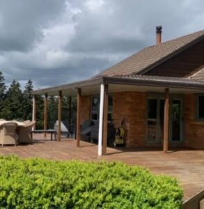 Brick house with a wide wooden deck and covered patio, tan wicker seating, under a cloudy sky.